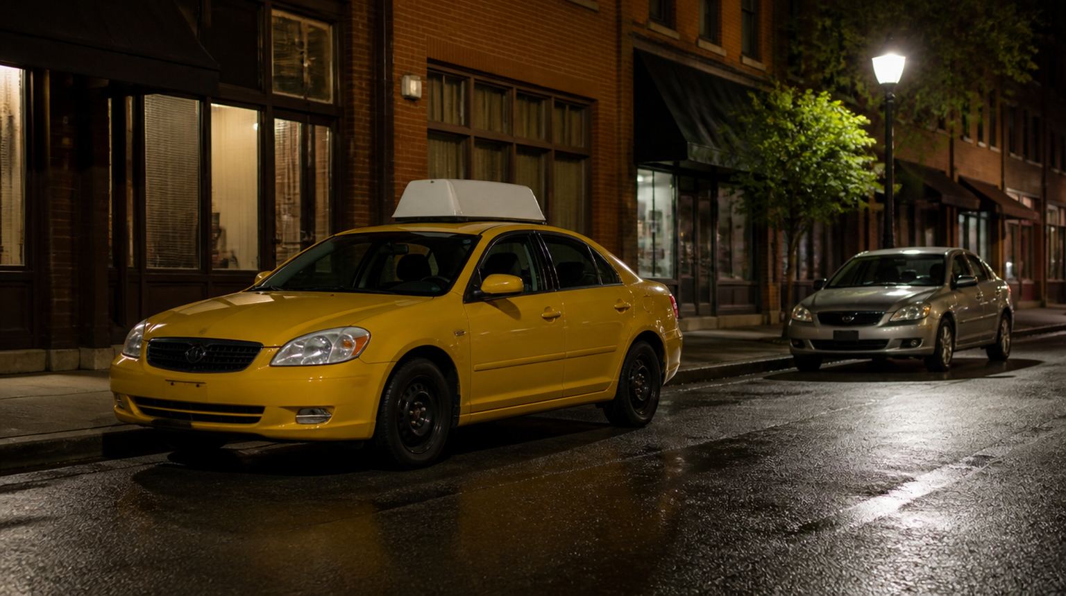 Yellow taxi parked at a city curb at night with an unmarked silver car further down the block